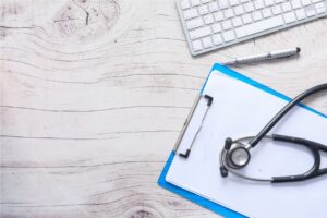 A stethoscope rests on a clipboard with papers, alongside a keyboard and pen, suggesting the review of Medical and Psychological Records.